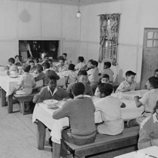 A black and white photograph of children at Kinchela Boys' Home. They are seated in the dining hall and eating a meal. 