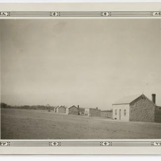 A black and white photograph of several buildings at Koonibba Lutheran Children's Home. The buildings are small, with stone-masonry and coarse plaster walls. Each building has two windows separated by a single door as well as a chimney.