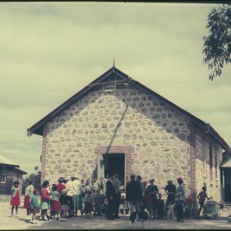 A colour photograph of a crowd at Marribank Mission. They are standing outside of a building with a stone-masonry façade. 