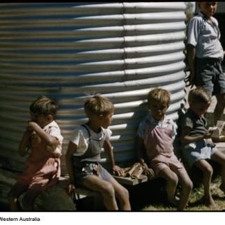 A colour photograph of a few young children at Marribank Mission. They are seated next to a water holding tank constructed from corrugated iron sheet metal. 