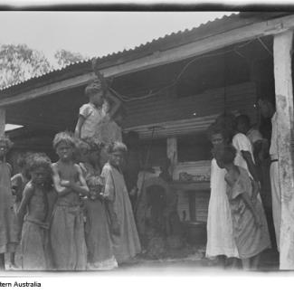 A black and white photograph of a group of people at Moore River Native Settlement. They are standing underneath a shelter made out of corrugated iron.
