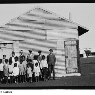 A black and white photograph of a group of people at Moore River Native Settlement. The children are all standing in front of a building which is made out of corrugated iron sheeting. The children are assembled together, with staff standing in the back row, and they are all are preparing for their photograph to be taken.