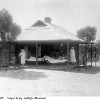 A black and white photograph of a medical ward at Moore River Native Settlement. There are a group of people lying in bed on the porch. There are staff members standing near them.