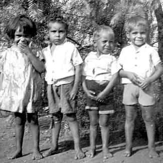 A black and white photograph of young children at Mount Margaret Mission. They are standing side-to-side in a row. The child on the left is hiding a laugh.