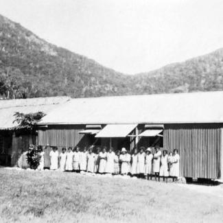 A black and white photograph people at Palm Island Dormitory. They are standing side-to-side in a row. They are outside and in front of a building constructed of corrugated iron sheet metal.