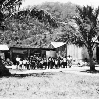 A black and white photograph of people at Palm Island Dormitory. They are standing side-to-side in front of a building constructed of corrugated iron sheet metal. There are two palm trees, one at either side of the group.
