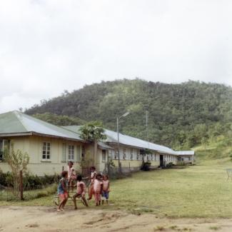 A colour photograph of a small group of children at Palm Island Dormitory. They are playing in front of the Girls' Dormitories, which are nestled at the foot of a hill that is dense with vegetation.