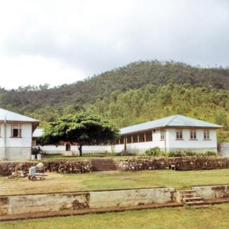 A colour photograph of the Boys' Dormitories at Palm Island Dormitory. The buildings are at the foot of a hill that is dense with vegetation, and a large tree is growing in the middle of the dormitories.