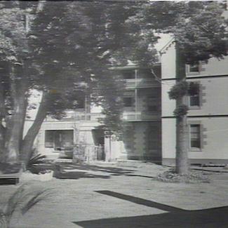 A black and white photograph of Parramatta Girls' Industrial School. There are two large trees growing in front of the building, and their foliage is shading benches beneath them.