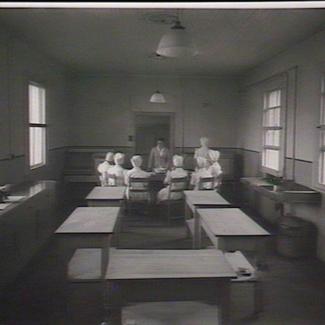 A black and white photograph of several staff members inside a room at Parramatta Girls' Industrial School. They are seated around a circular table and are listening to one person who is standing up and speaking. 