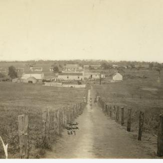 A sepia-toned photograph of Point McLeay Mission. A narrow, unpaved track is leading down to the Mission. The mission consists of several small buildings clustered together in a large, open field.