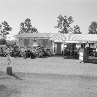 A black and white photograph of a crowd at Retta Dixon Home. They are seated outside in the open are are listening to a person speaking at a podium.