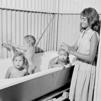 A black and white photograph of a small group of children at Retta Dixon Home. Three younger children are in a bath, while an older child is assisting them.
