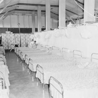 A black and white photograph of the sleeping quarters at Retta Dixon Home. Arranged in rows are a large number of metal framed beds. A single person is fitting sheets over the beds.