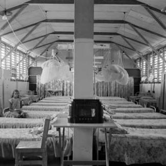 A black and white photograph of the sleeping quarters at Retta Dixon Home. Arranged in rows are a large number of metal framed beds. A single person is seated on one bed.