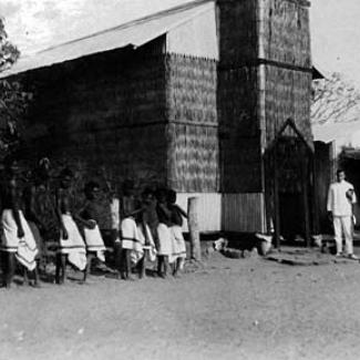 A black and white photograph of a group of people at Roper River Mission. People, comprising both residents and staff members, are assembled outside the Mission church. 
