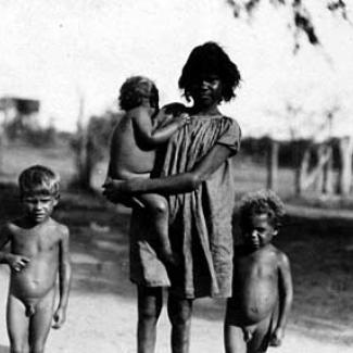 A black and white photograph of an adult and young children at Roper River Mission. The adult is holding one child, while the other two children are walking. 