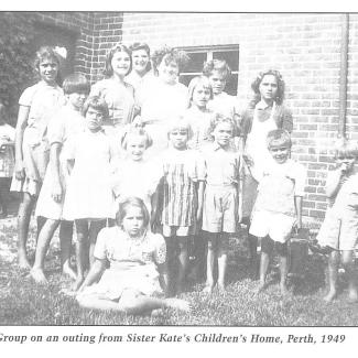 A black and white photograph of a group of children from Sister Kate's Children's Home. They are assembled together and preparing to be photographed. They are standing on the grass outside of a brick-faced building.