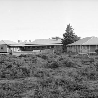 A black and white photograph of several buildings at Umeewarra Mission Children's Home. A chain link fence surroundings the buildings, while beyond the fence is a grassy plain.