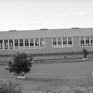 A black and white photograph of a building at Umeewarra Mission Children's Home. The building is separated from the road by a chain link fence. The building itself is long, with most of its length covered by window. Inside the windows hang cards that spell: Umeewarra School.