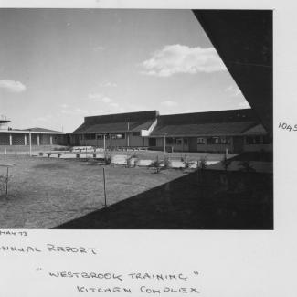 A black and white photograph of Westbrook Training Centre. On the photograph is written: May seventythree, Annual Report, Westbrook Training, Kitchen Complex. The kitchen building opens out into a large grassy space with several saplings.