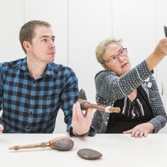 A man holds an ancient stone hatchet while the woman next to him examines a stone axe-head. More examples of ancient tools rest on the table infront of them.