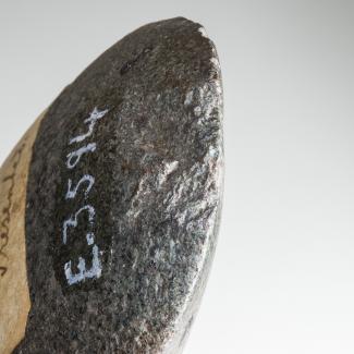 A close-up photograph of an ancient stone axe-head, lightly chipped at the edge and labelled in white paint with a cataloging number.